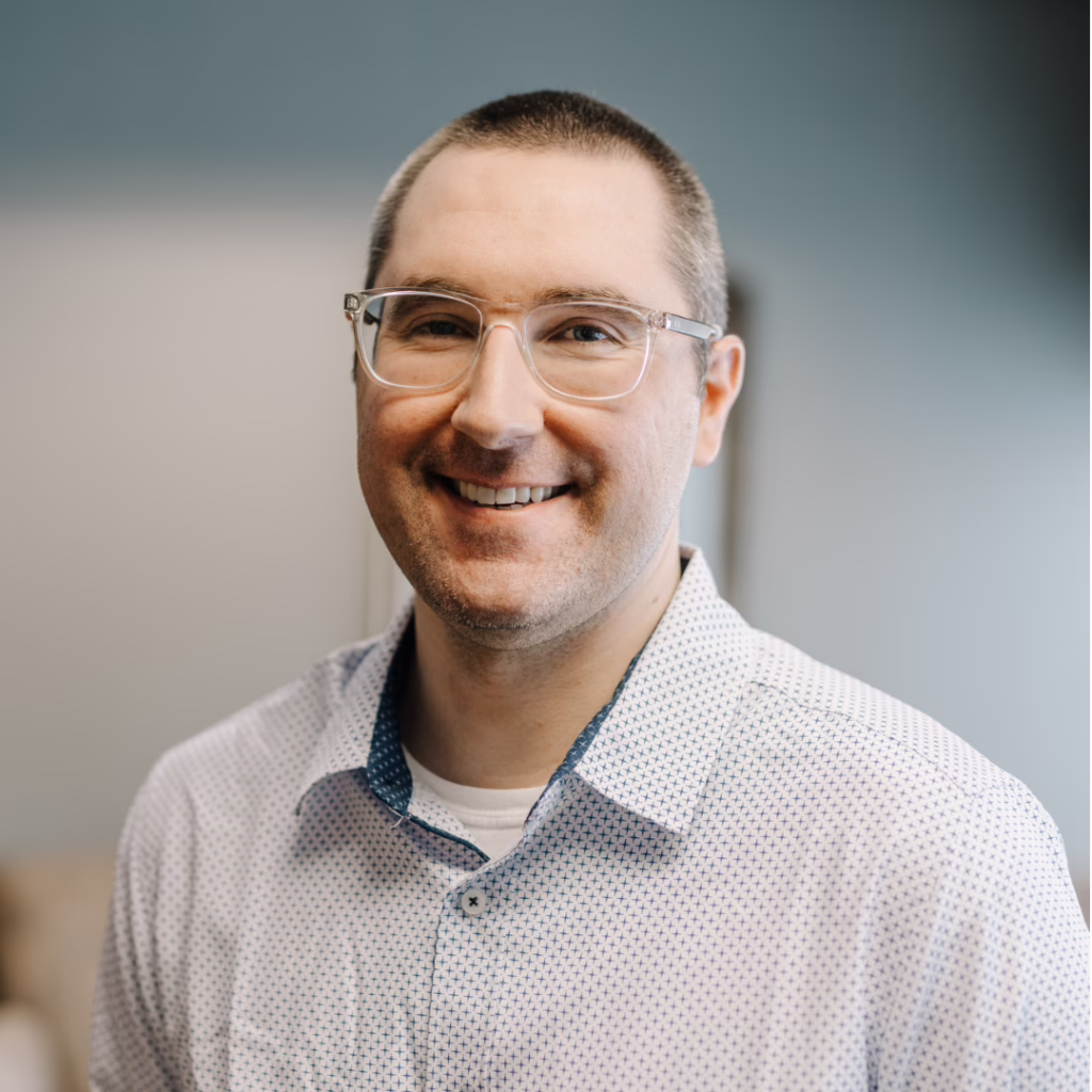 Head-and-shoulders portrait of a smiling man with clear-framed glasses wearing a light patterned shirt, looking at the camera while indoors.