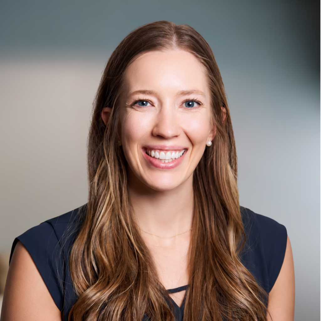Head-and-shoulders portrait of a smiling woman with long light brown hair in a navy sleeveless top, neutral background behind her.